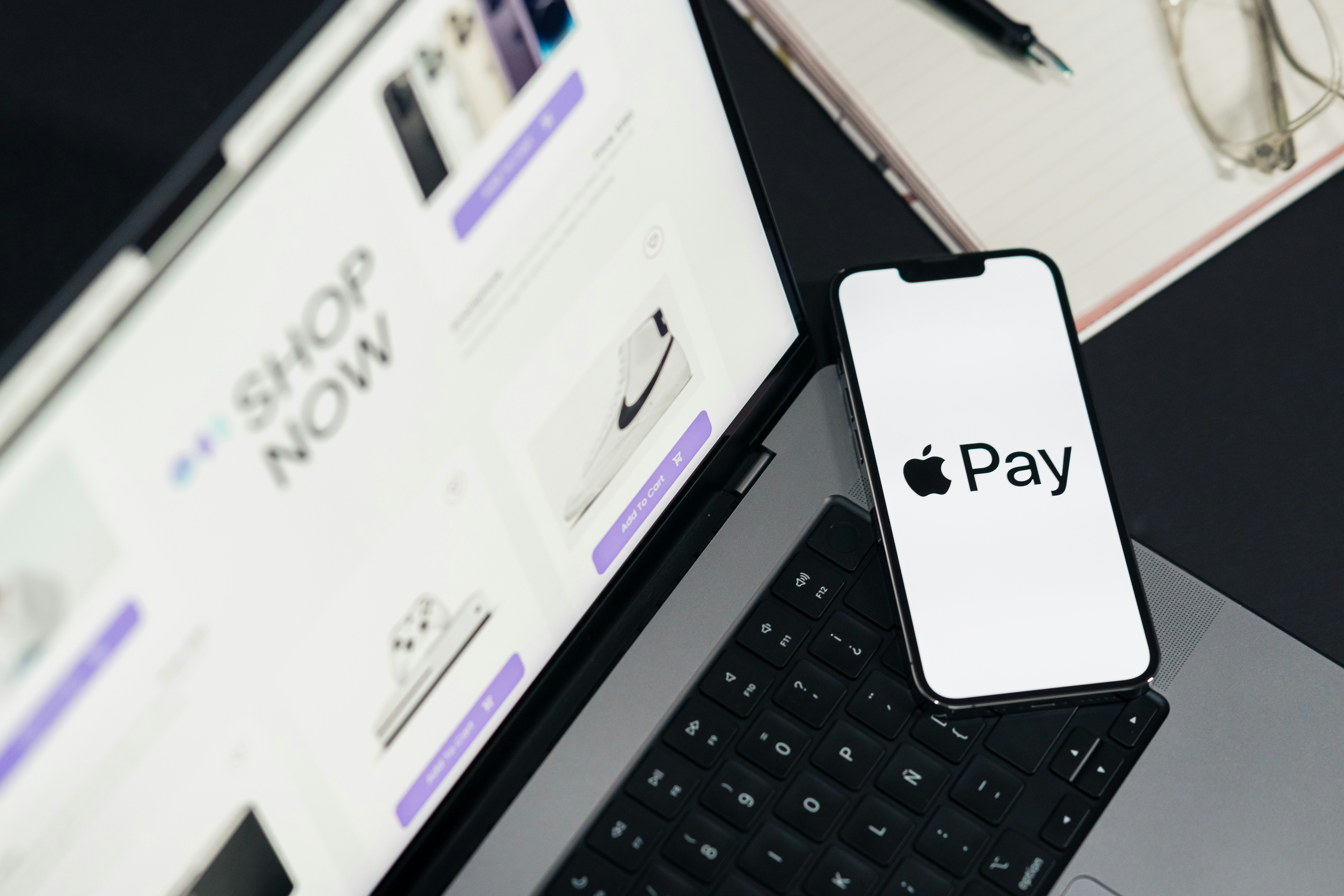 Apple Pay on phone screen. Top view of workspace. Table desk with laptop with ecommerce on screen, mobile, notepad